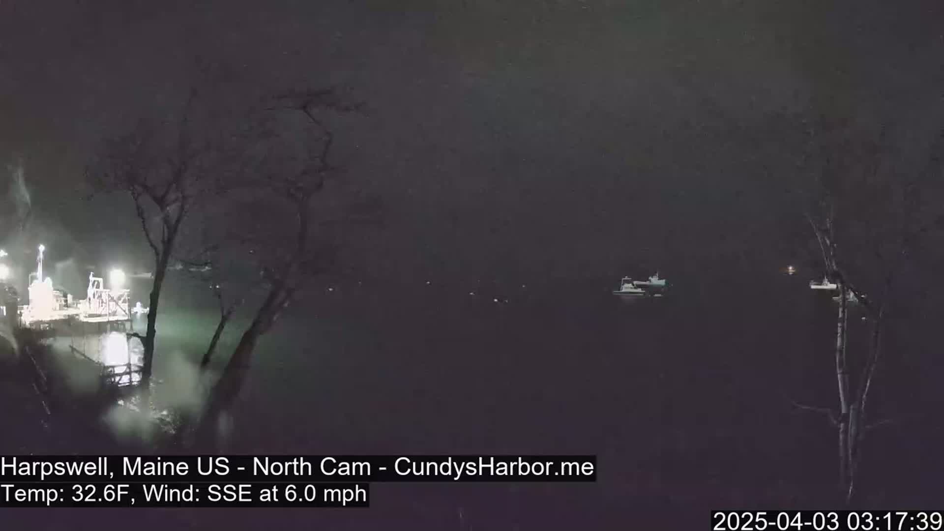 A nighttime view of a dark body of water with several boats and a dock, under snowy conditions with a 6.0 mph wind from the SSE.
