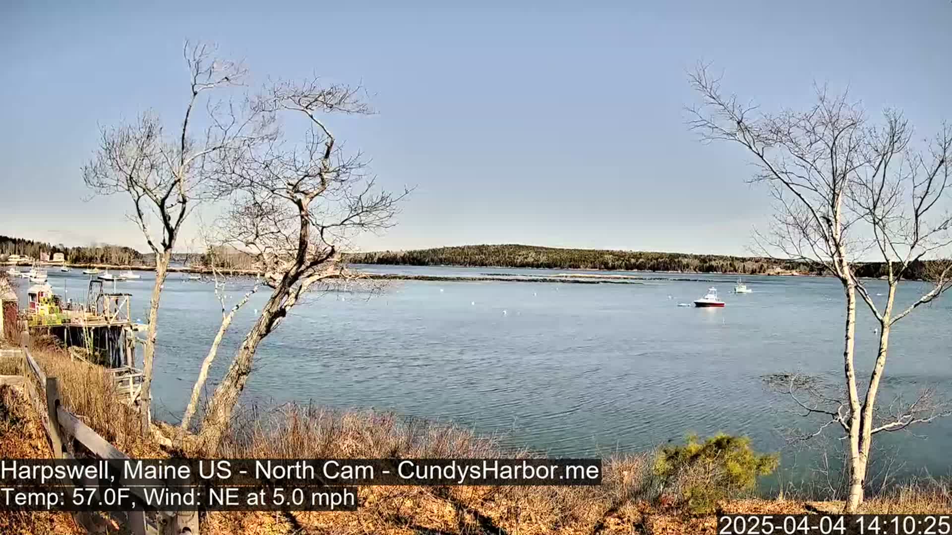 A calm body of water with a few boats is seen from a shoreline with bare trees and a partly sunny sky.