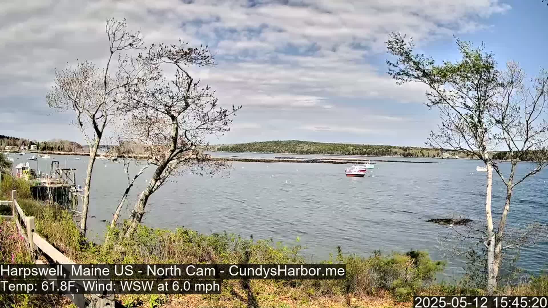 A partly cloudy day with a 6 mph wind shows a calm bay with several small boats near a rocky shoreline, partially obscured by bare and budding trees.