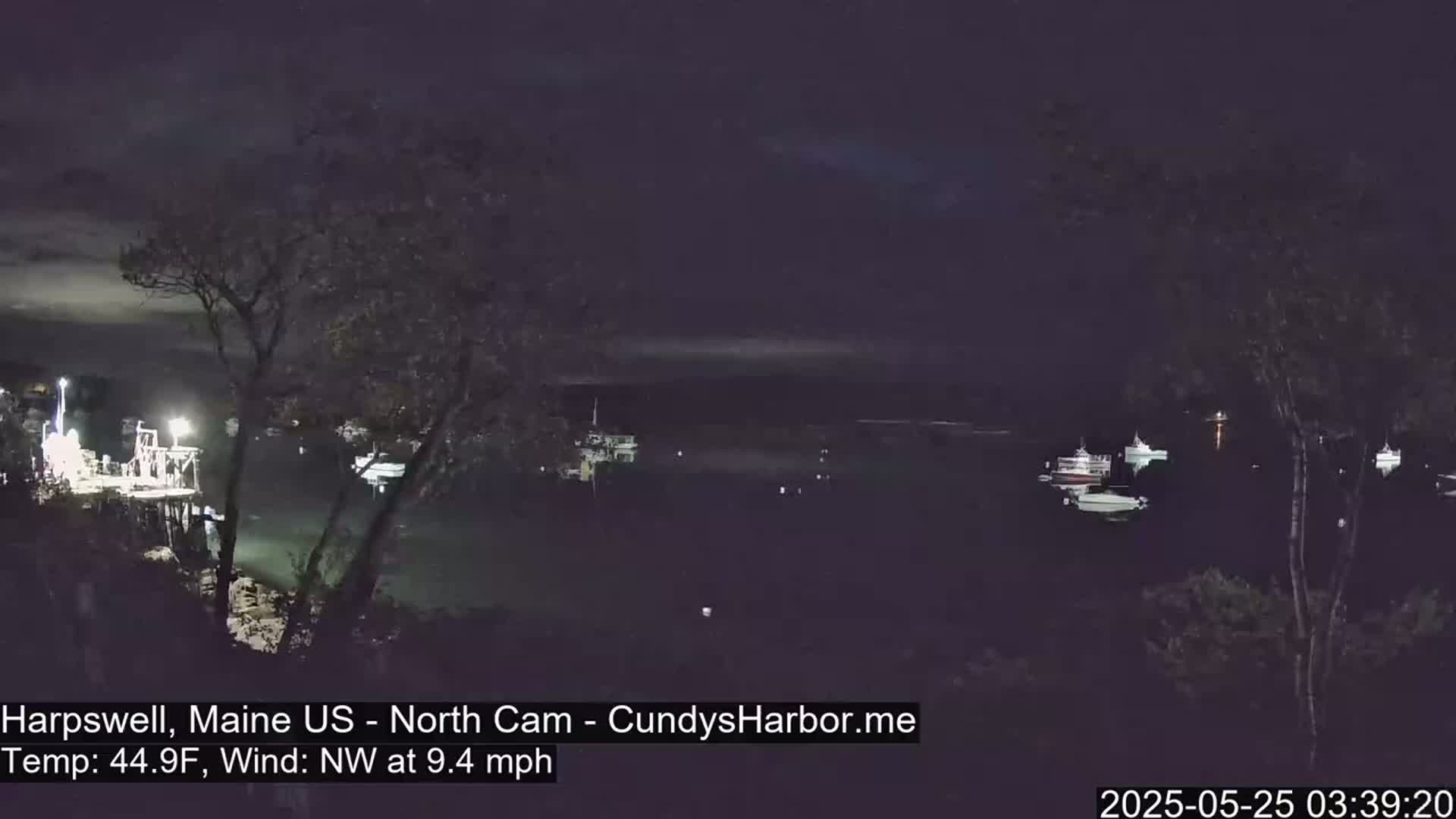 A nighttime view of a harbor with several boats moored, under a dark, mostly clear sky, with a northwest wind.