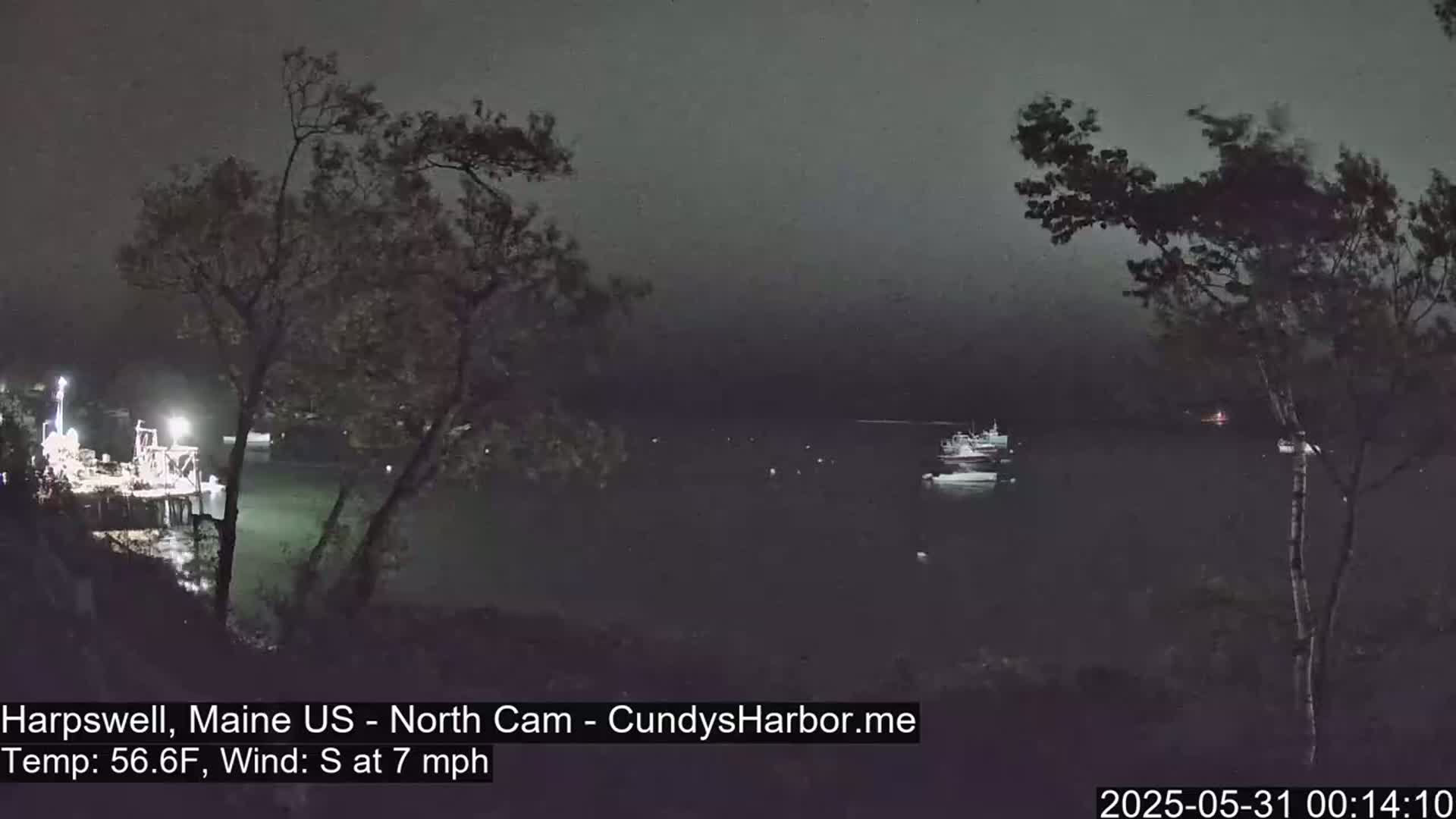 A nighttime view of a dark body of water with several boats, lit structures on the shore, and trees silhouetted against a hazy sky under a 7 mph south wind.