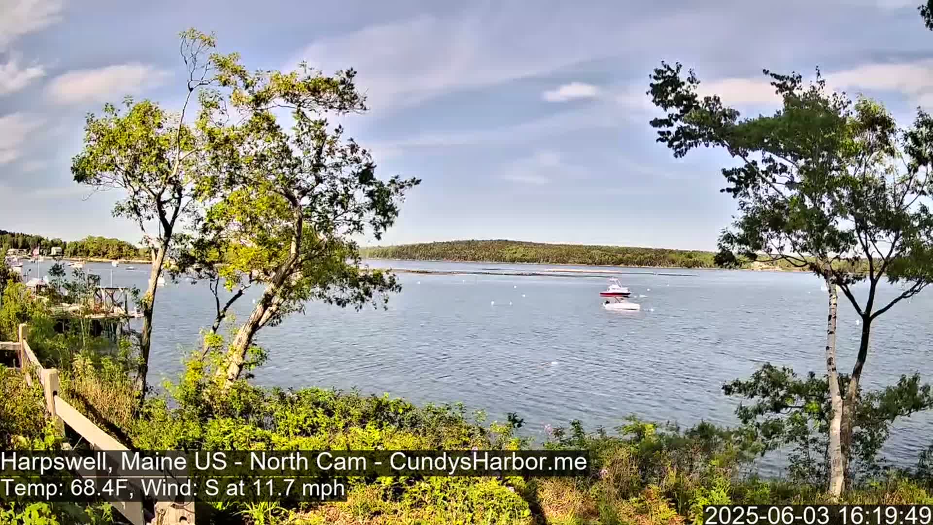 A tranquil harbor scene under a partly cloudy sky shows a calm body of water with a small boat, and leafy green trees in the foreground.