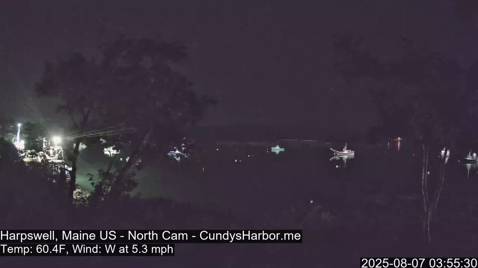 A nighttime view of a harbor with several boats, under a dark sky with a west wind at 5.3 mph and a temperature of 60.4°F.