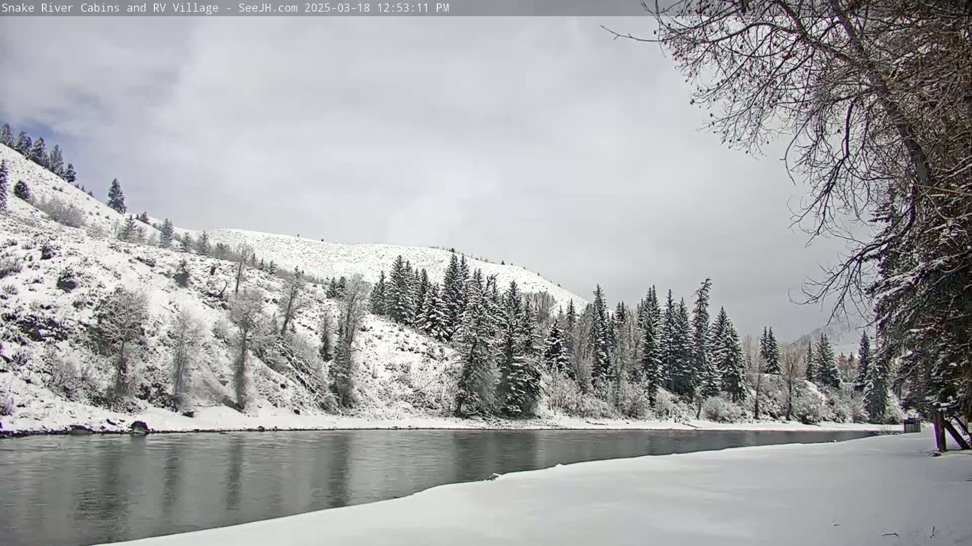 A snow-covered landscape featuring a calm river reflecting the overcast sky, with snow-laden hills and evergreen trees lining the riverbanks.
