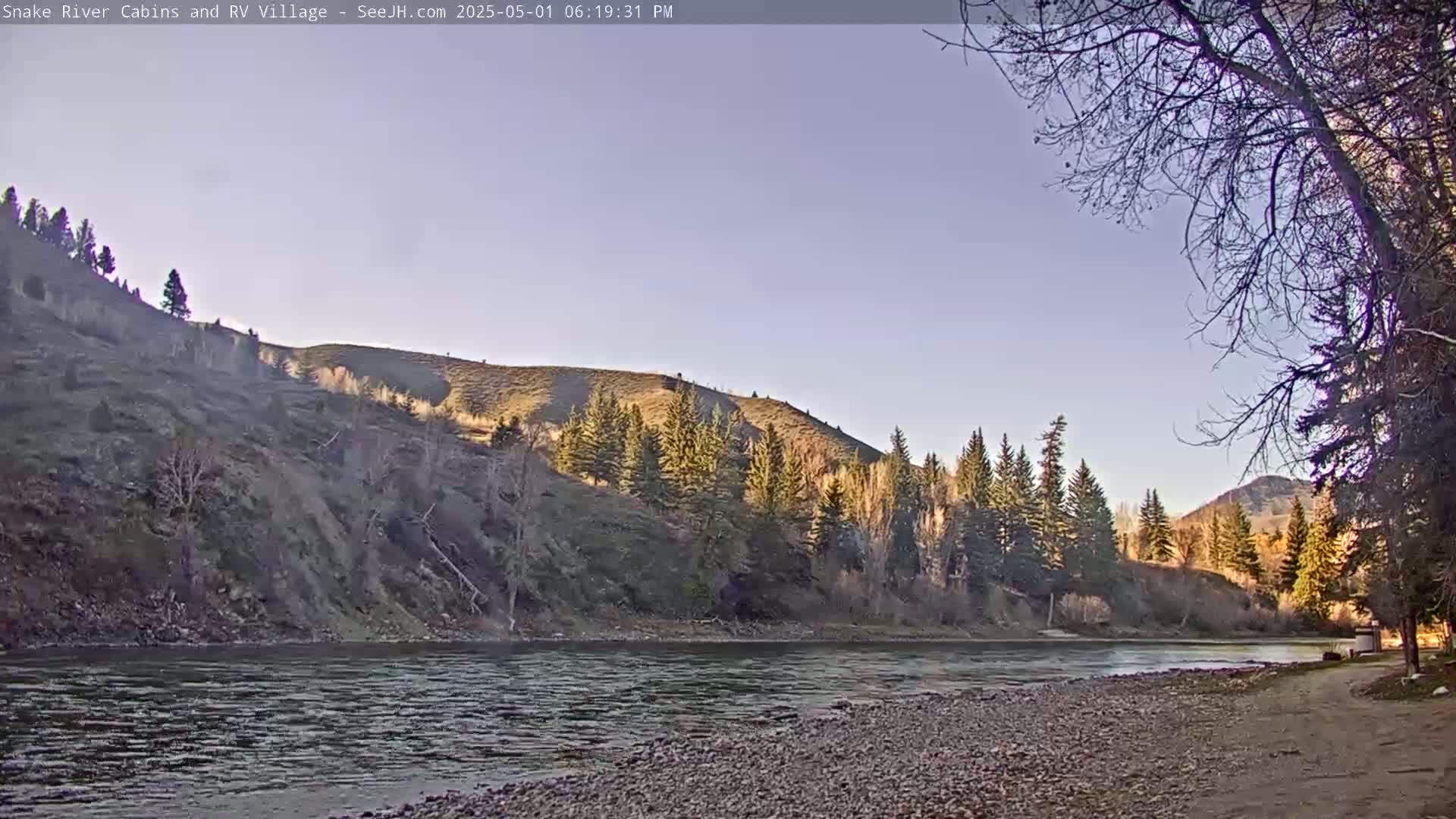 A calm river flows past a rocky shore, under a clear, pale blue sky, with hills covered in sparse, late-autumn trees in the background.