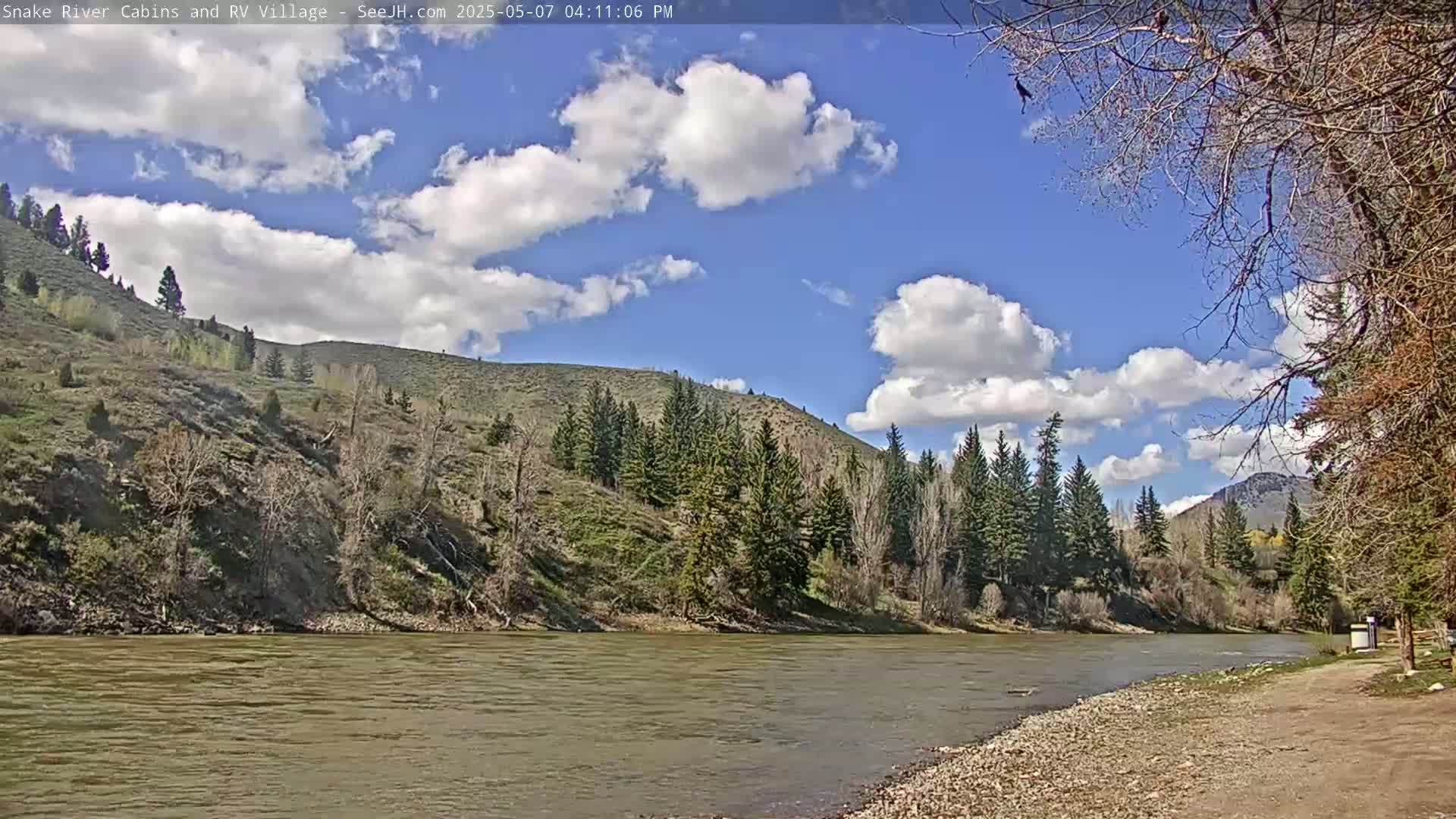 A wide, muddy river flows under a partly cloudy blue sky past a rocky shore and hills covered in sparse trees and scrub.