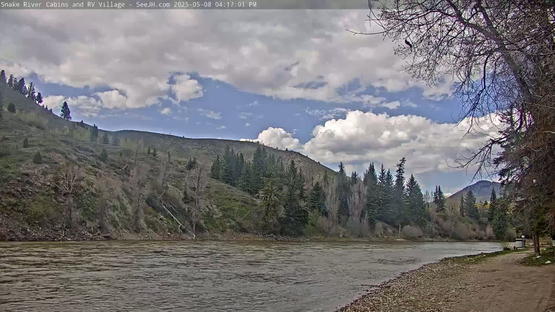 A partly cloudy day reveals a river flowing past a rocky, tree-lined shoreline with hills in the background.