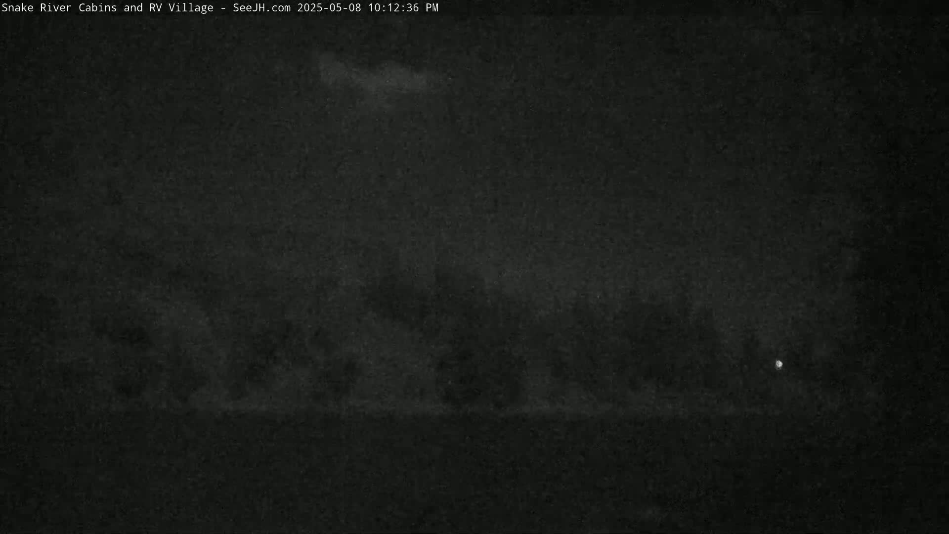 A nighttime, low-light image shows a dark, indistinct landscape with a few faint lights visible in the distance.