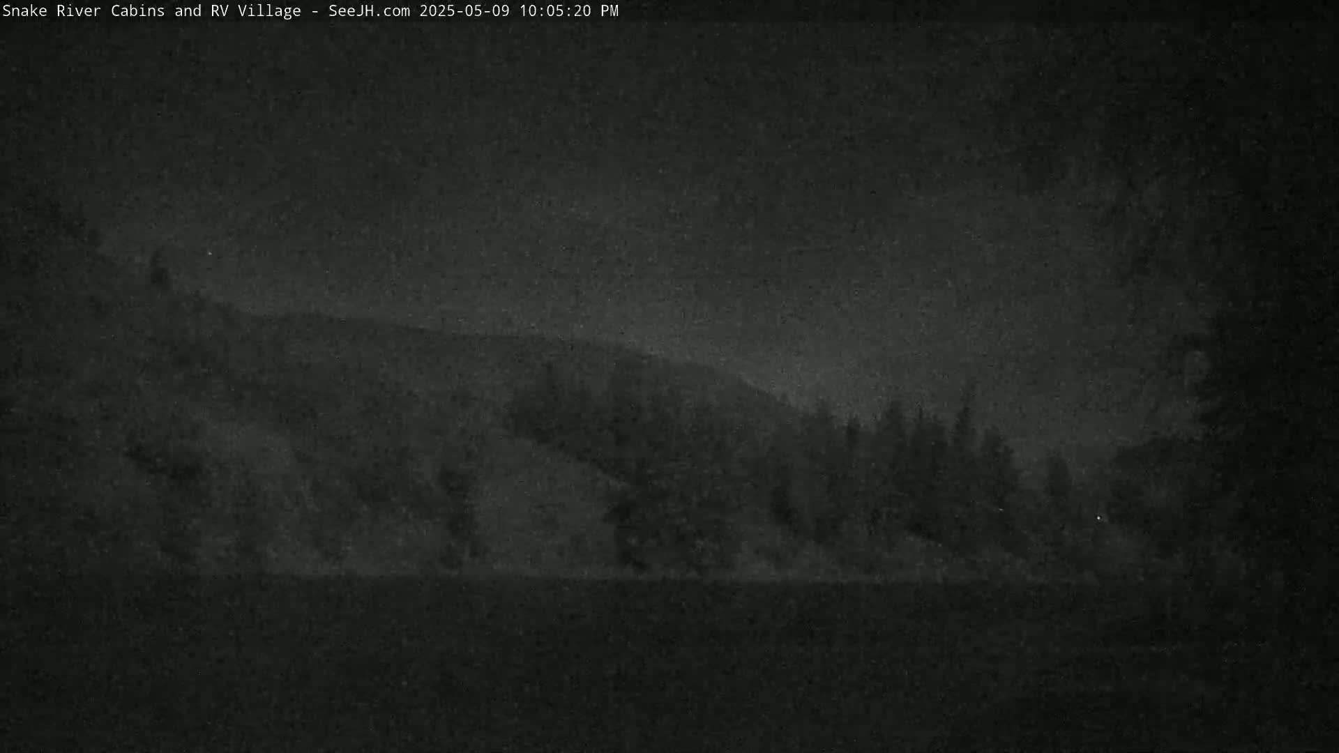 A nighttime, low-light view shows a dark hill or mountain silhouetted against a dark sky, with a line of dark trees at the base of the hill.