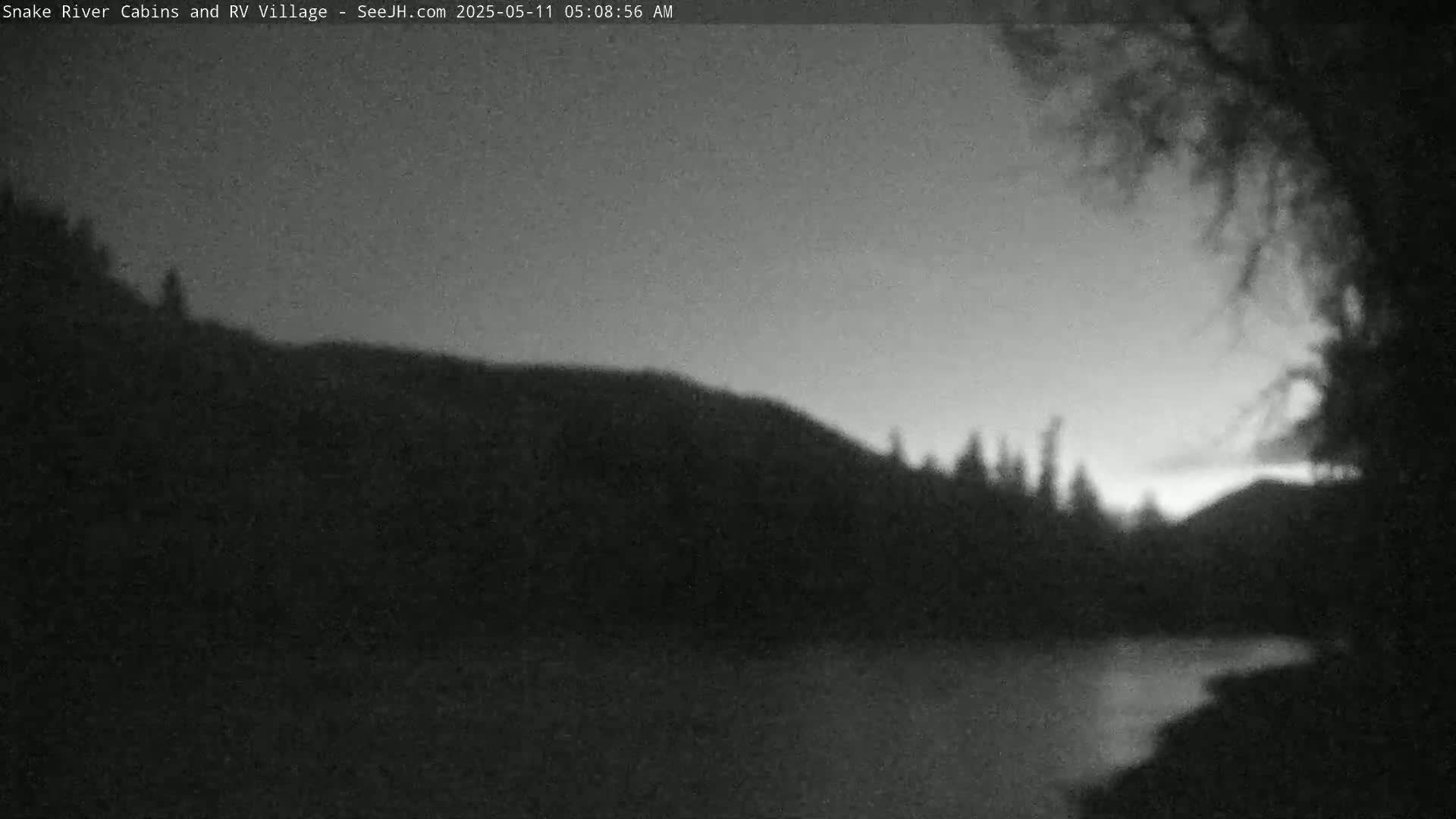 A dark, pre-dawn scene shows a calm body of water reflecting a dusky sky, bordered by silhouetted trees and hills.
