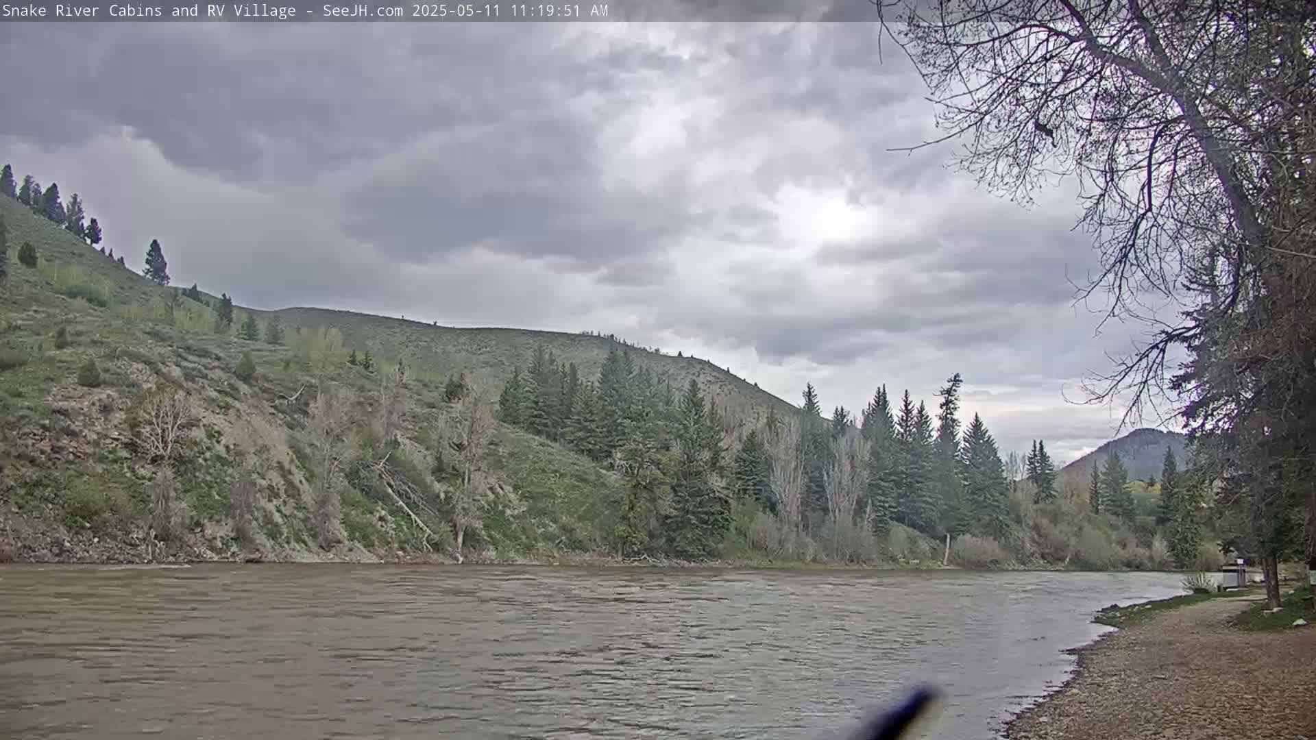 A wide, calm river flows under a cloudy sky past a rocky, tree-covered hillside.