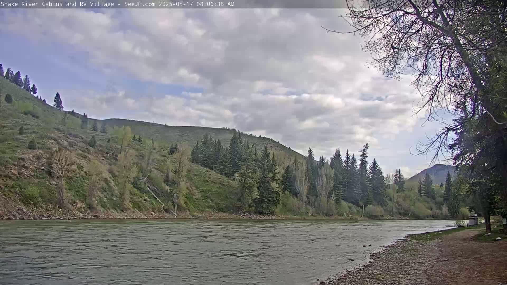 A partly cloudy day reveals a wide river flowing past a grassy riverbank, with hills covered in evergreens and deciduous trees in the background.