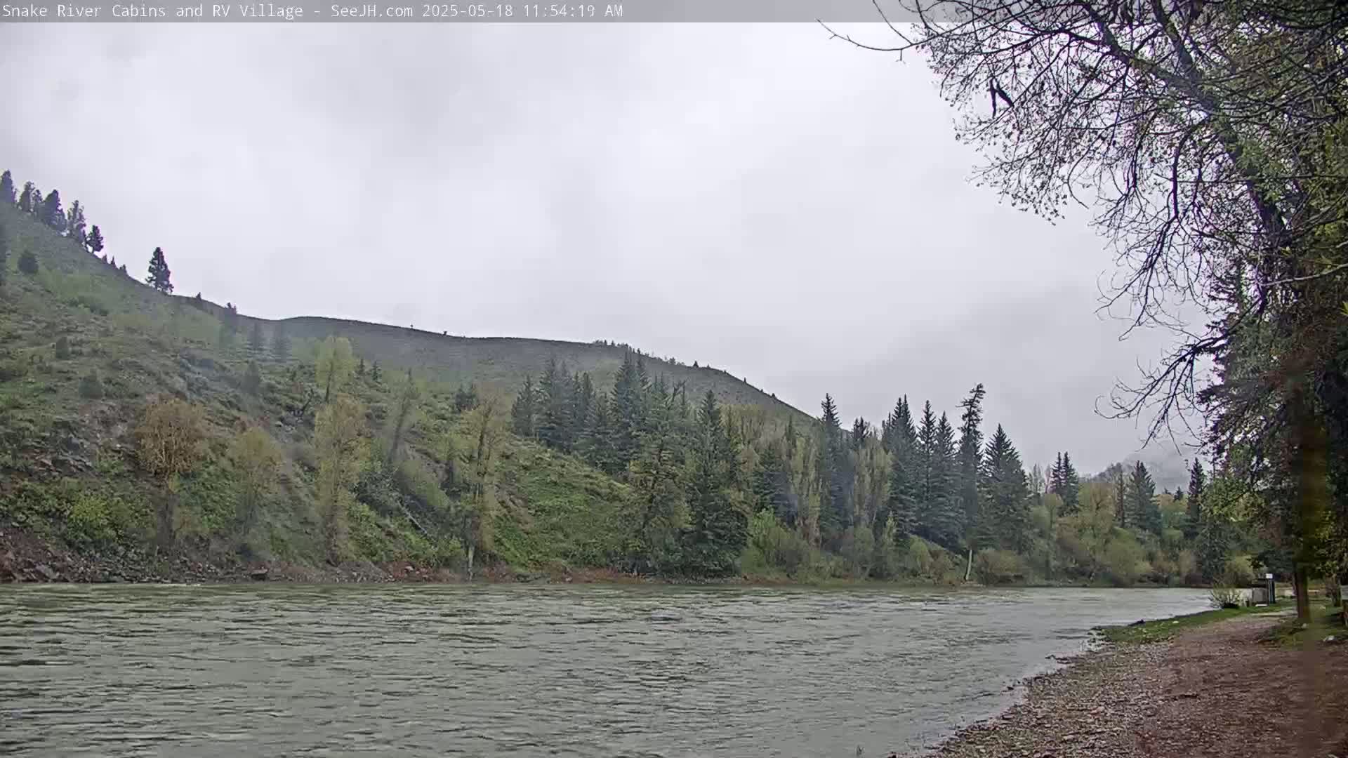 A wide, grey river flows beneath an overcast sky past a rocky, tree-covered hillside.