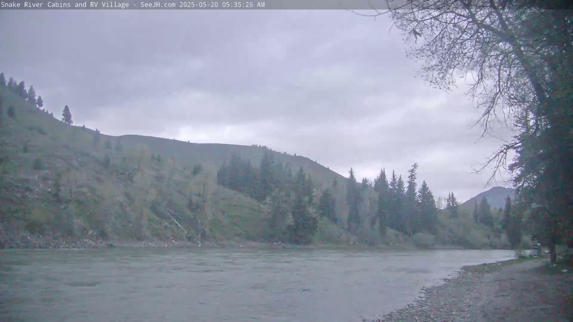 A wide, grey river flows beneath an overcast sky past a gently sloping hillside covered in sparse trees.