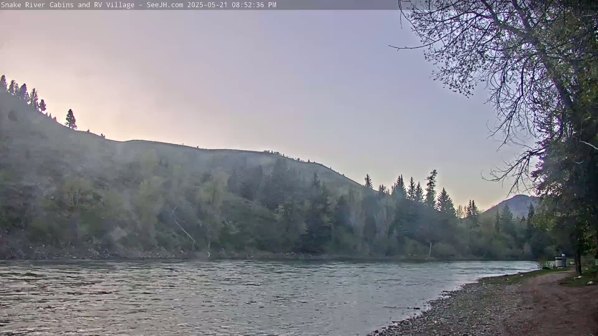 A calm river flows past a rocky, tree-covered bank under a pale, dusk sky.
