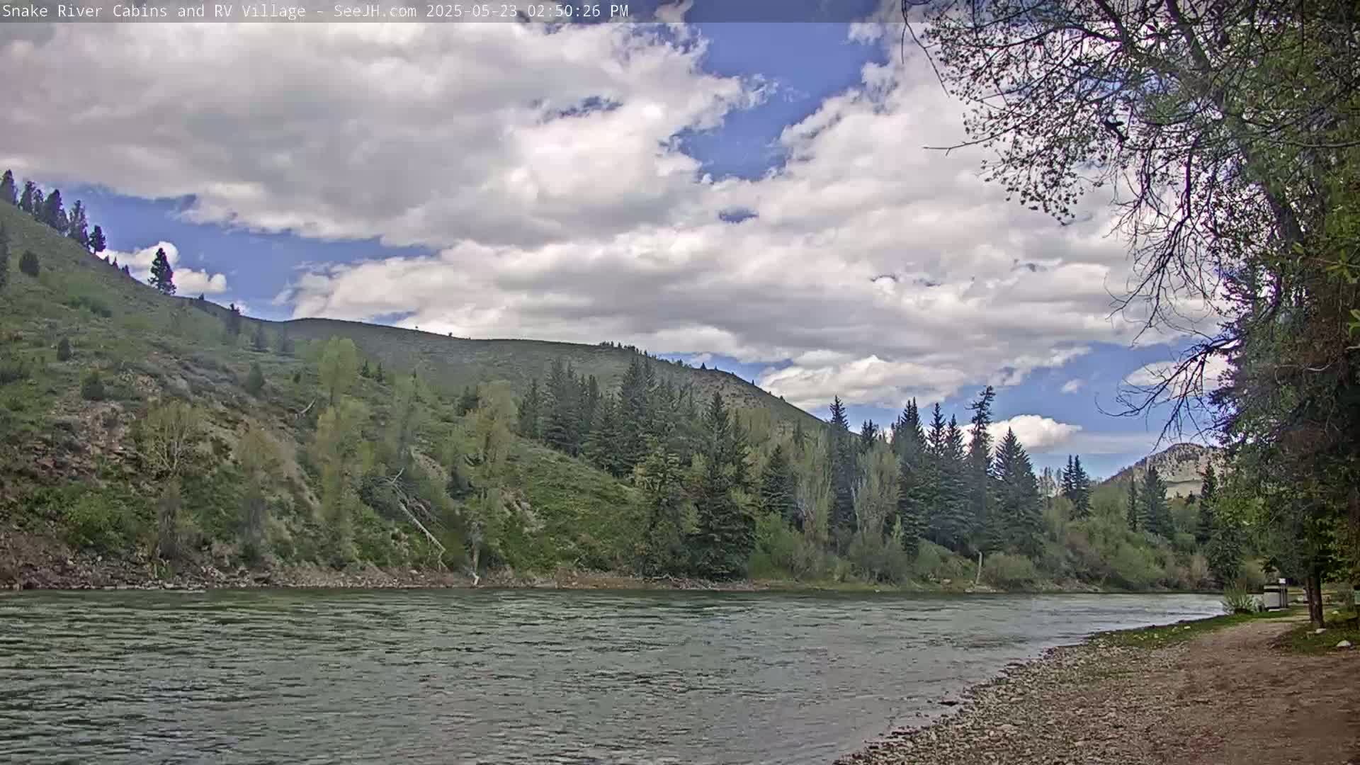 A wide river flows past a rocky bank under a partly cloudy sky, with lush green hills and evergreens rising in the background.