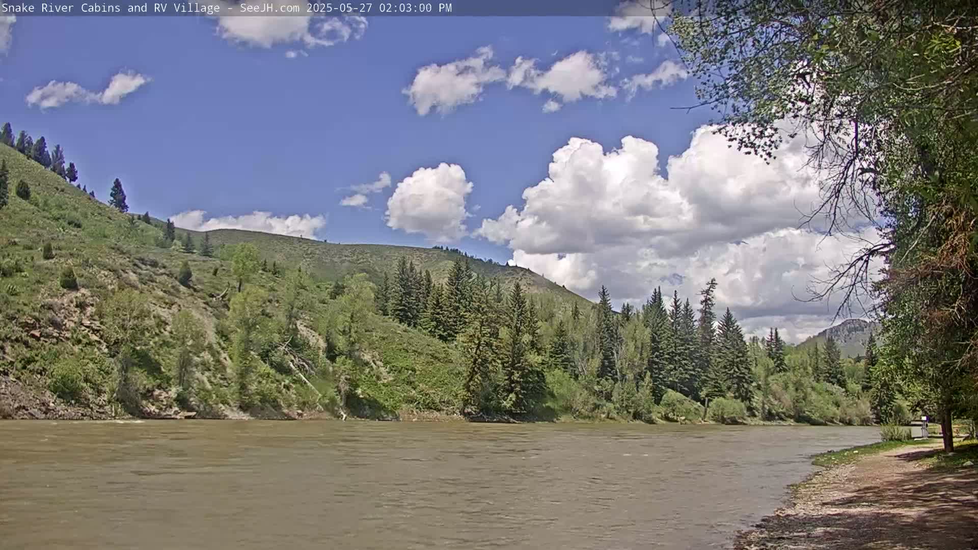 A wide, calm river flows past a rocky, tree-covered hillside under a partly cloudy blue sky.