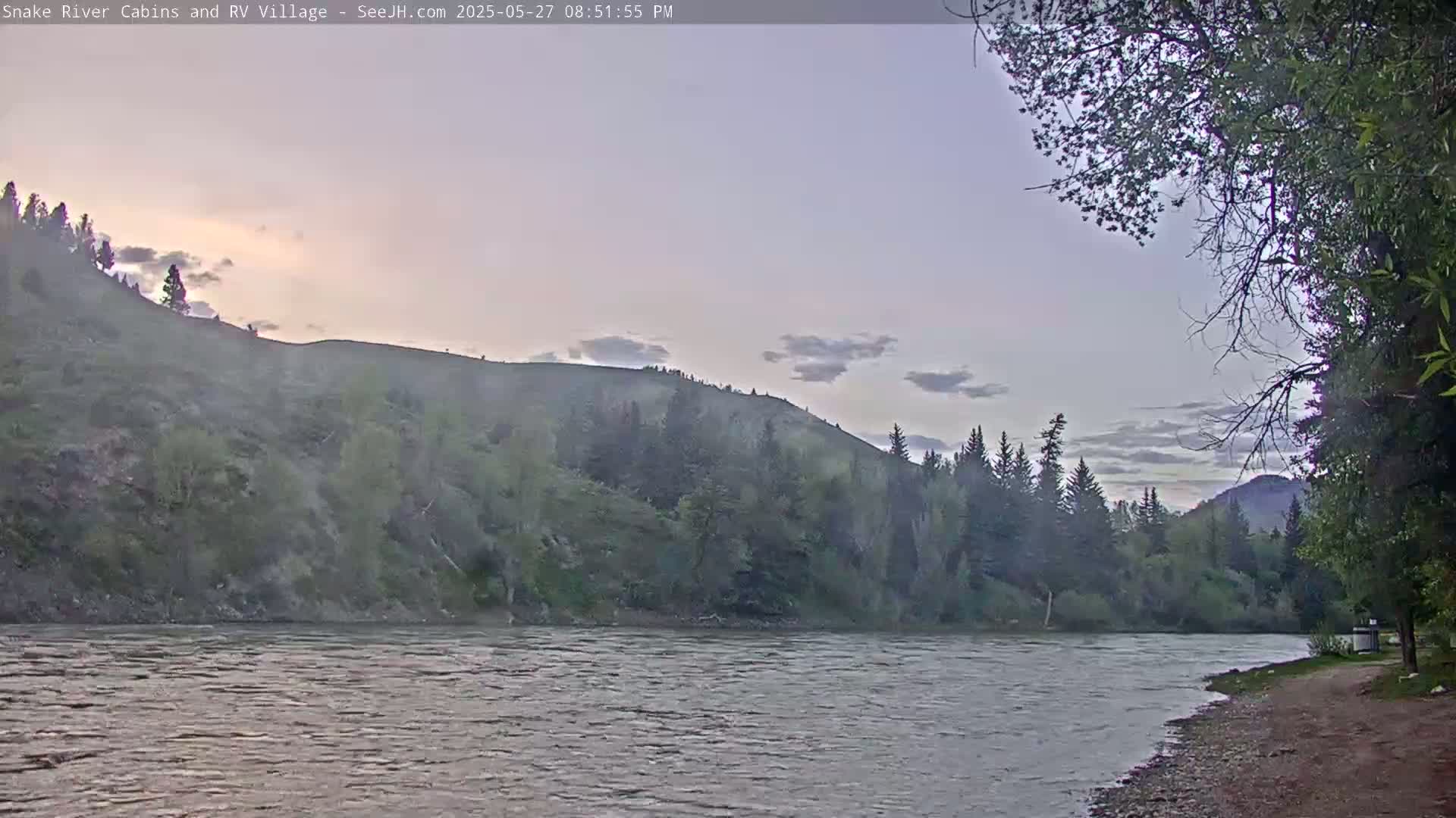A calm river flows past a tree-lined bank and a hill under a dusky, cloudy sky.