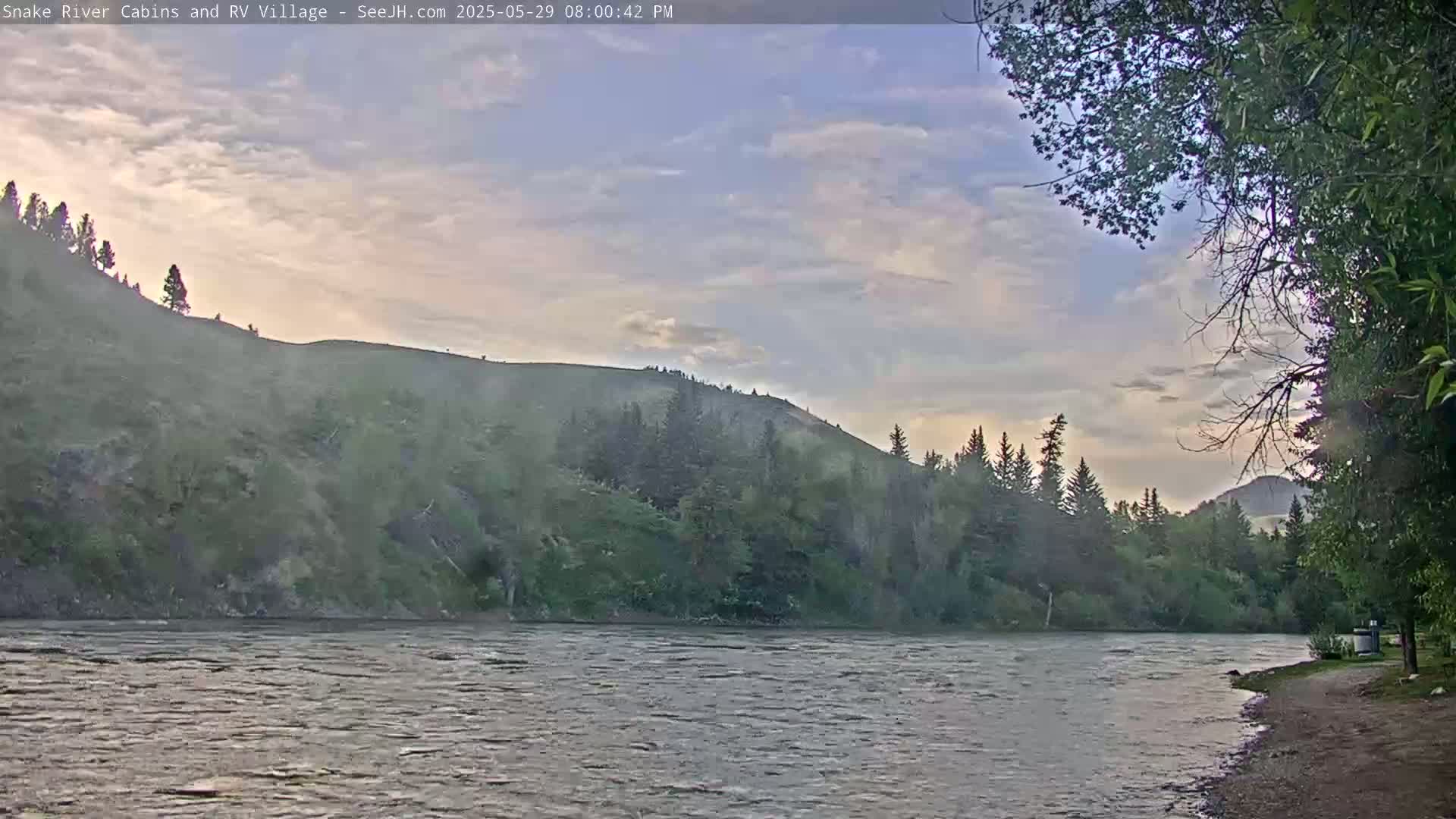 A partly cloudy evening sky overlooks a wide, gently flowing river bordered by a sandy bank and verdant hills covered in trees.