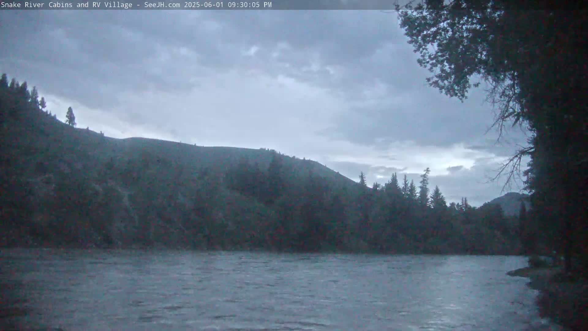 A wide, grey river flows beneath a cloudy, overcast sky past dark green hills and trees.