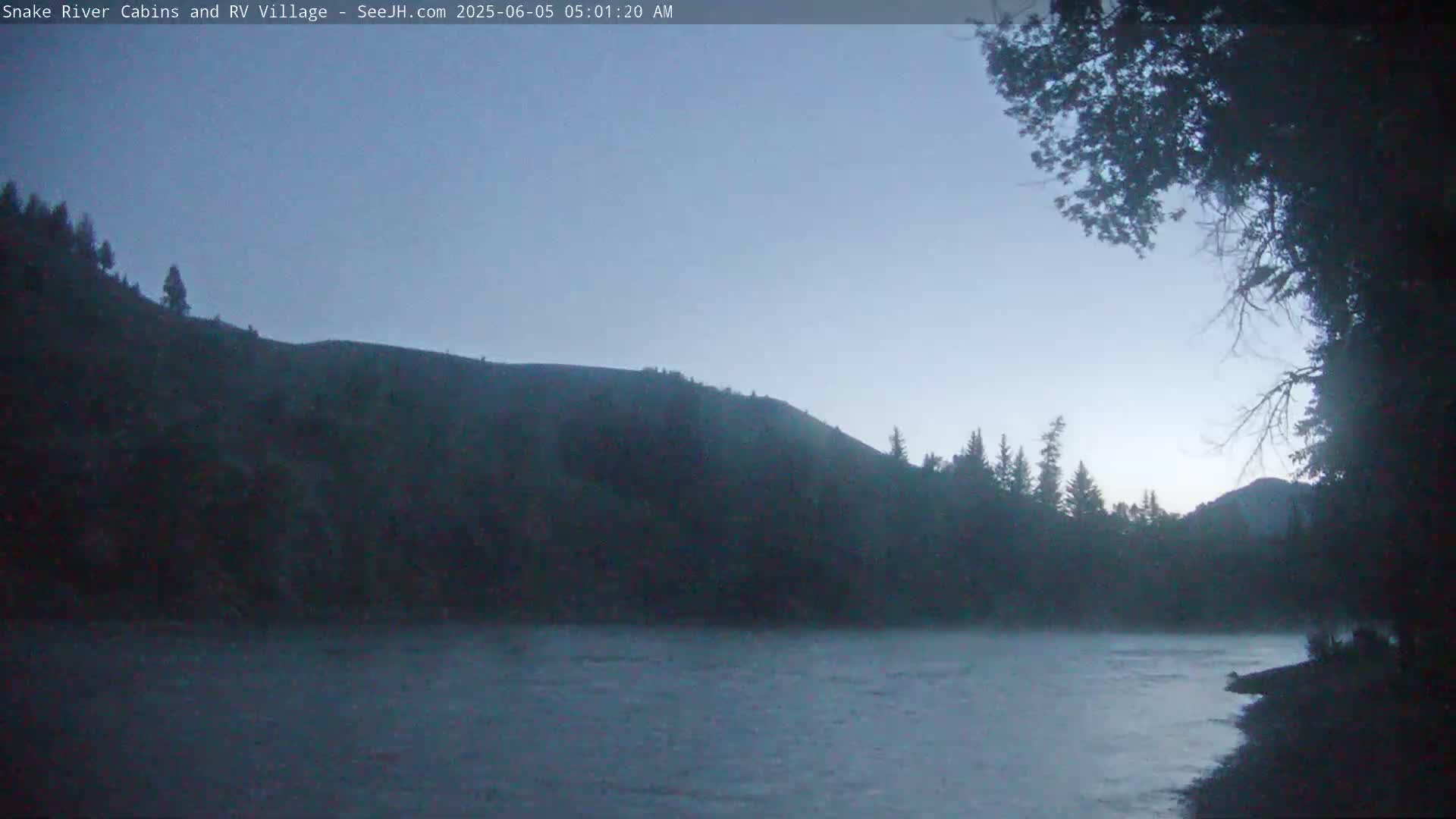 A dark, calm river flows beneath a dusky sky, bordered by silhouetted hills and trees.