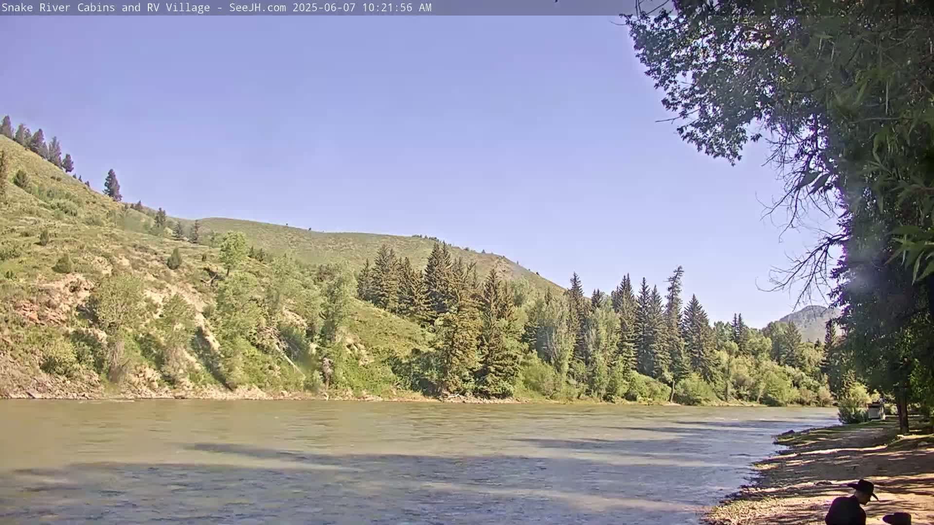 A wide, calm river flows past a sandy bank and lush green hills under a clear blue sky.