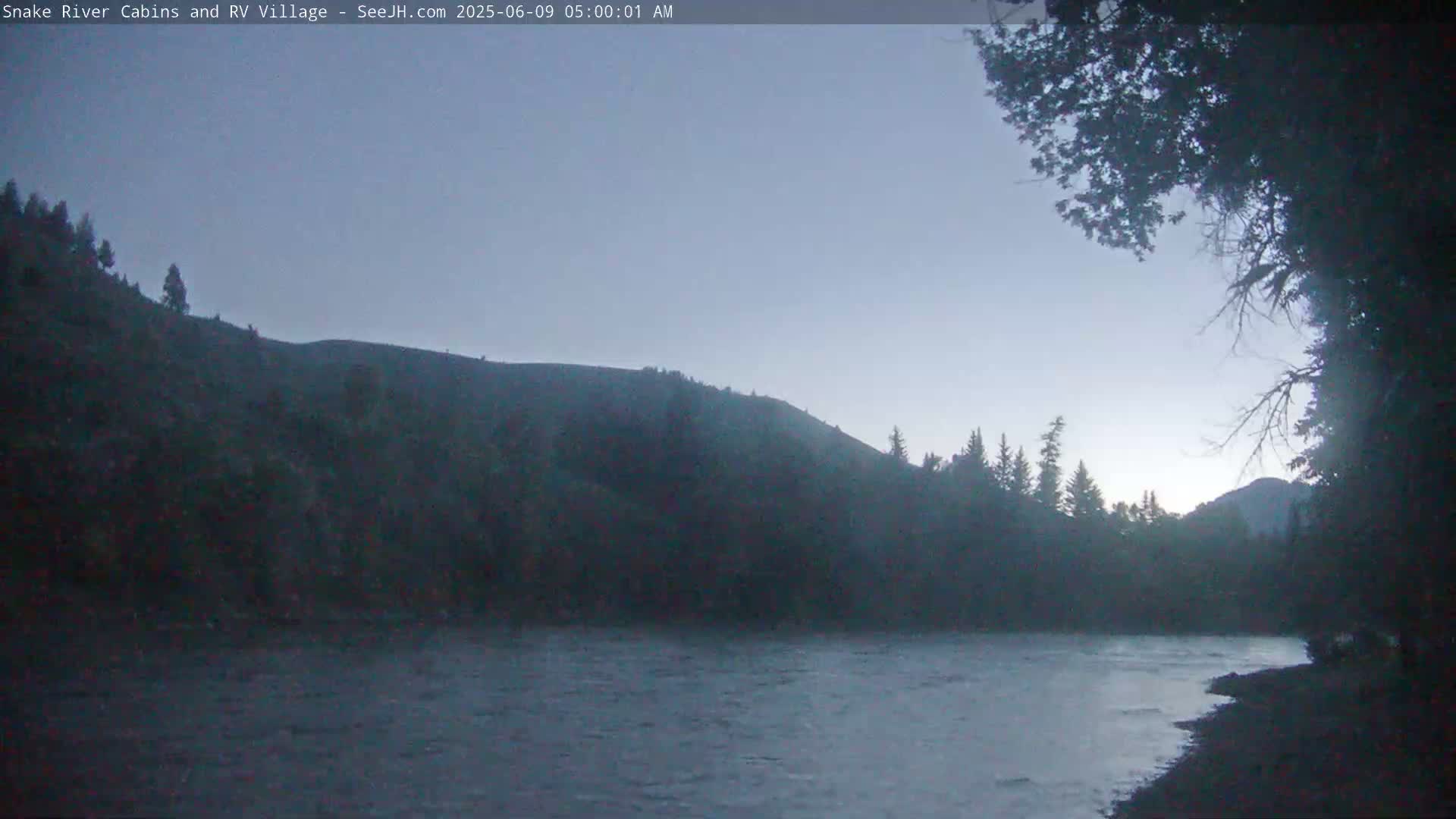 A calm river flows in front of a dark, tree-covered hill under a dusky, pre-dawn sky.