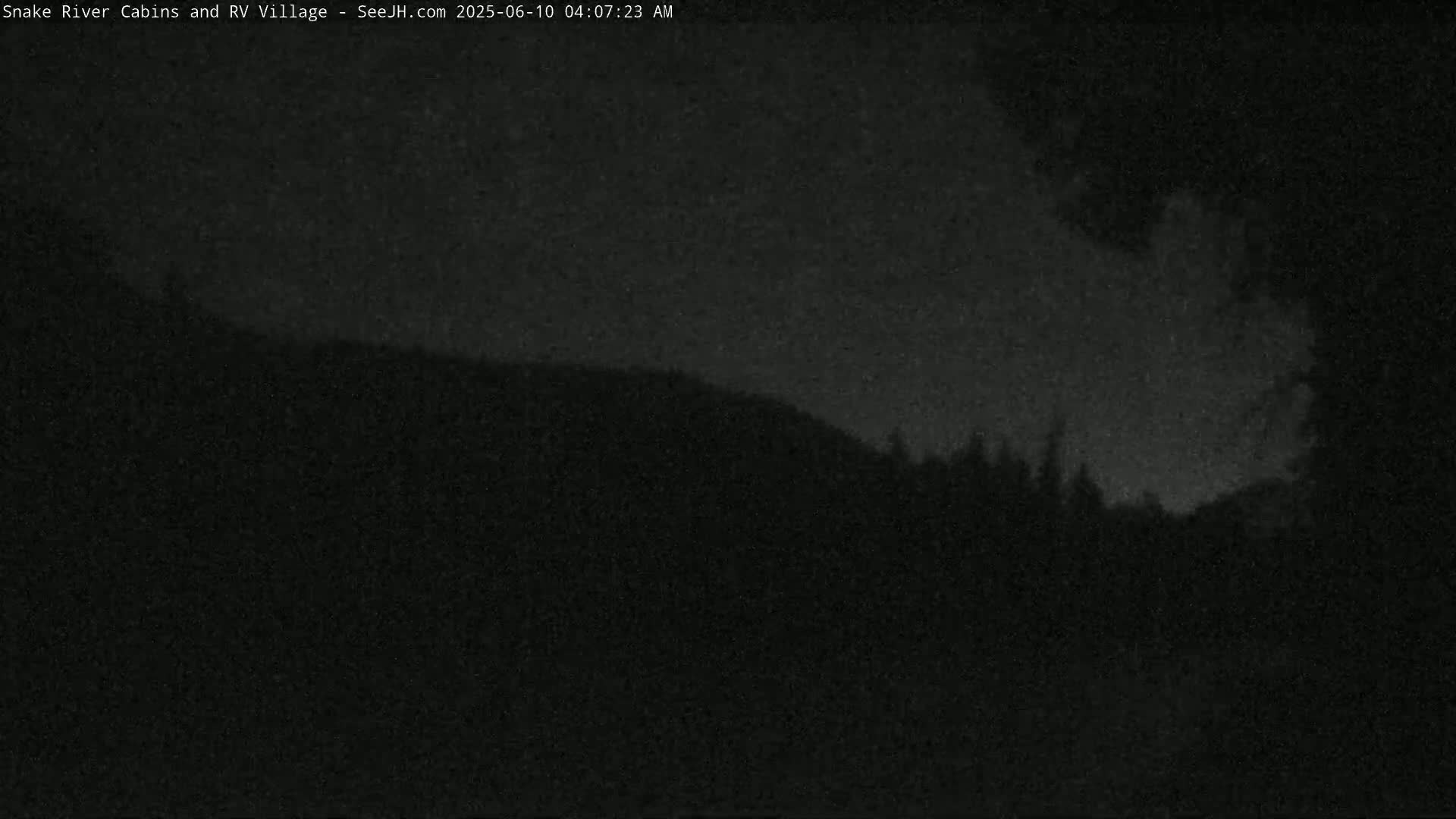 A nighttime, low-light view shows a dark, silhouetted hillside with a few trees under a mostly clear, dark sky.