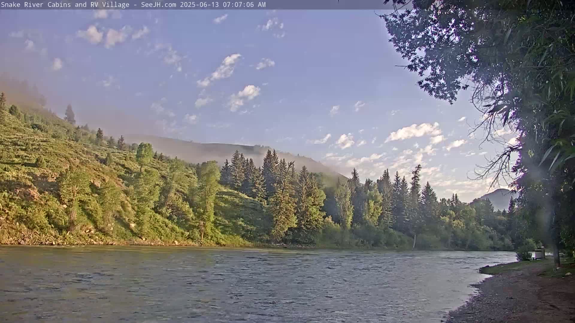 A calm river flows past a lush, green hillside covered in trees under a partly cloudy, clear sky.
