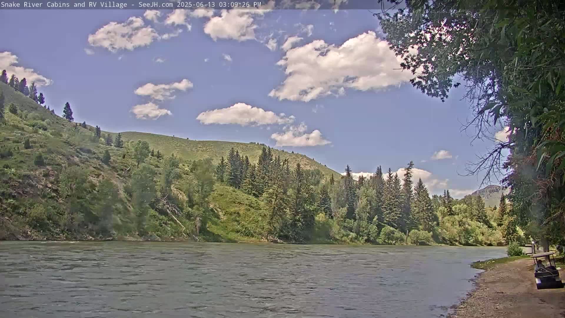 A partly cloudy sky looks over a calm river flowing beside a lush, green hillside covered in evergreen trees.