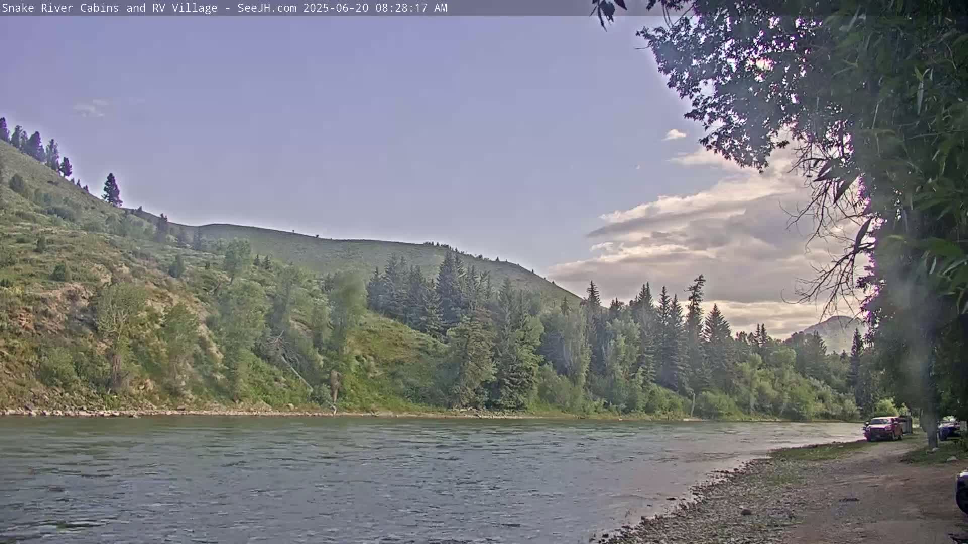 A partly cloudy sky overlooks a wide river flowing past a green, tree-covered hillside.