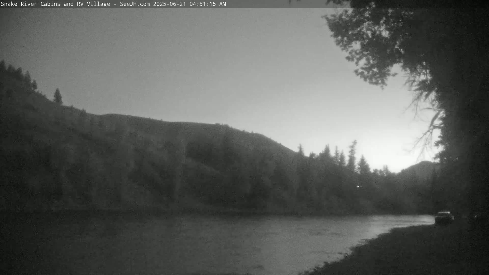 A dark, pre-dawn scene shows a calm river flowing past a wooded hillside under a light gray sky.