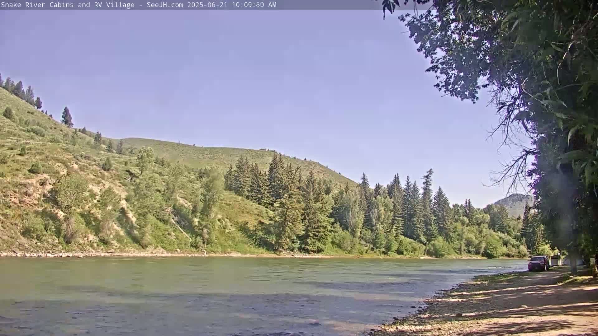 A calm river flows past a grassy bank lined with evergreen trees and hills under a clear blue sky.