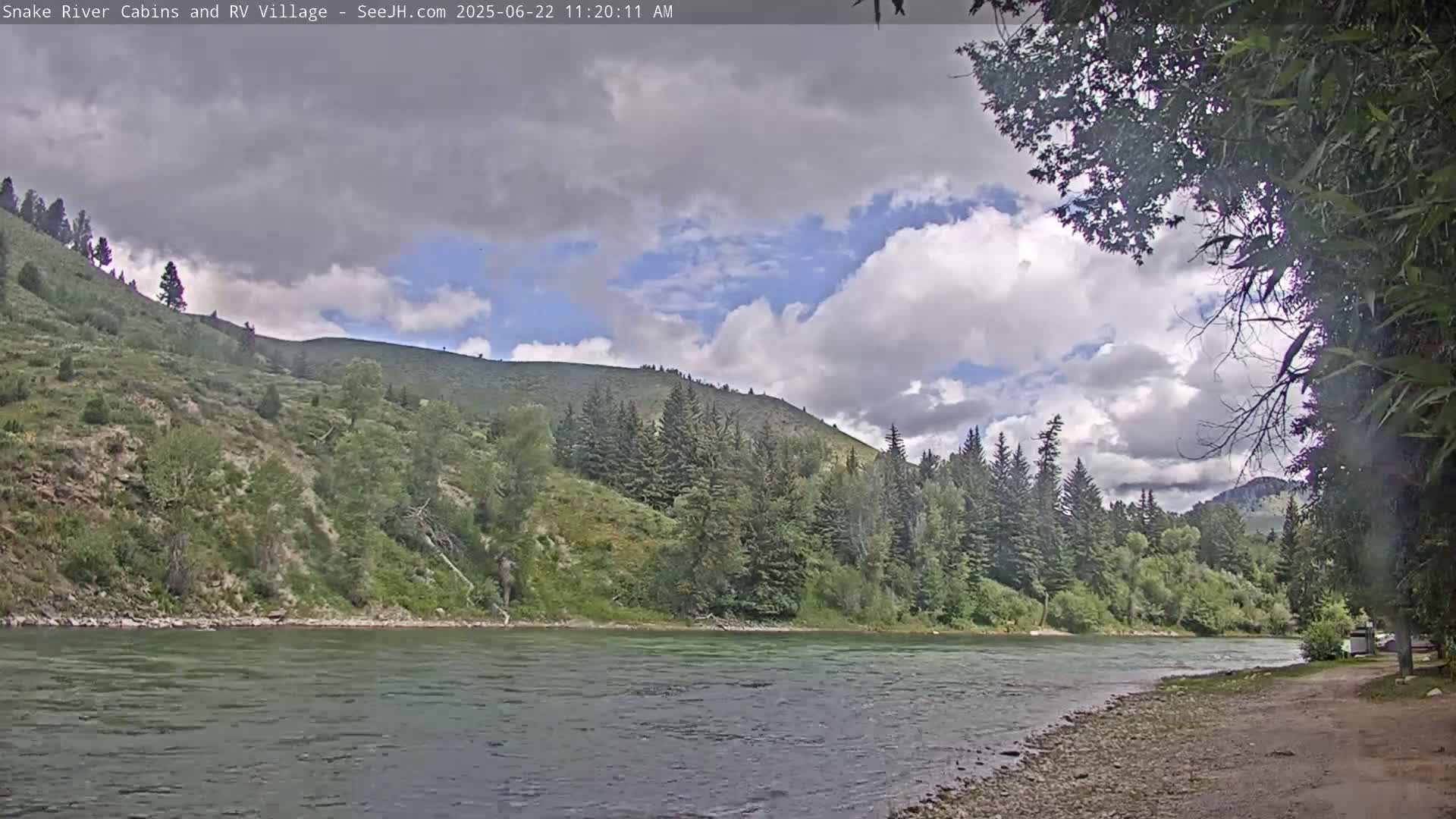 A partly cloudy day reveals a calm river flowing beside a rocky shore and verdant hills covered in evergreen trees.
