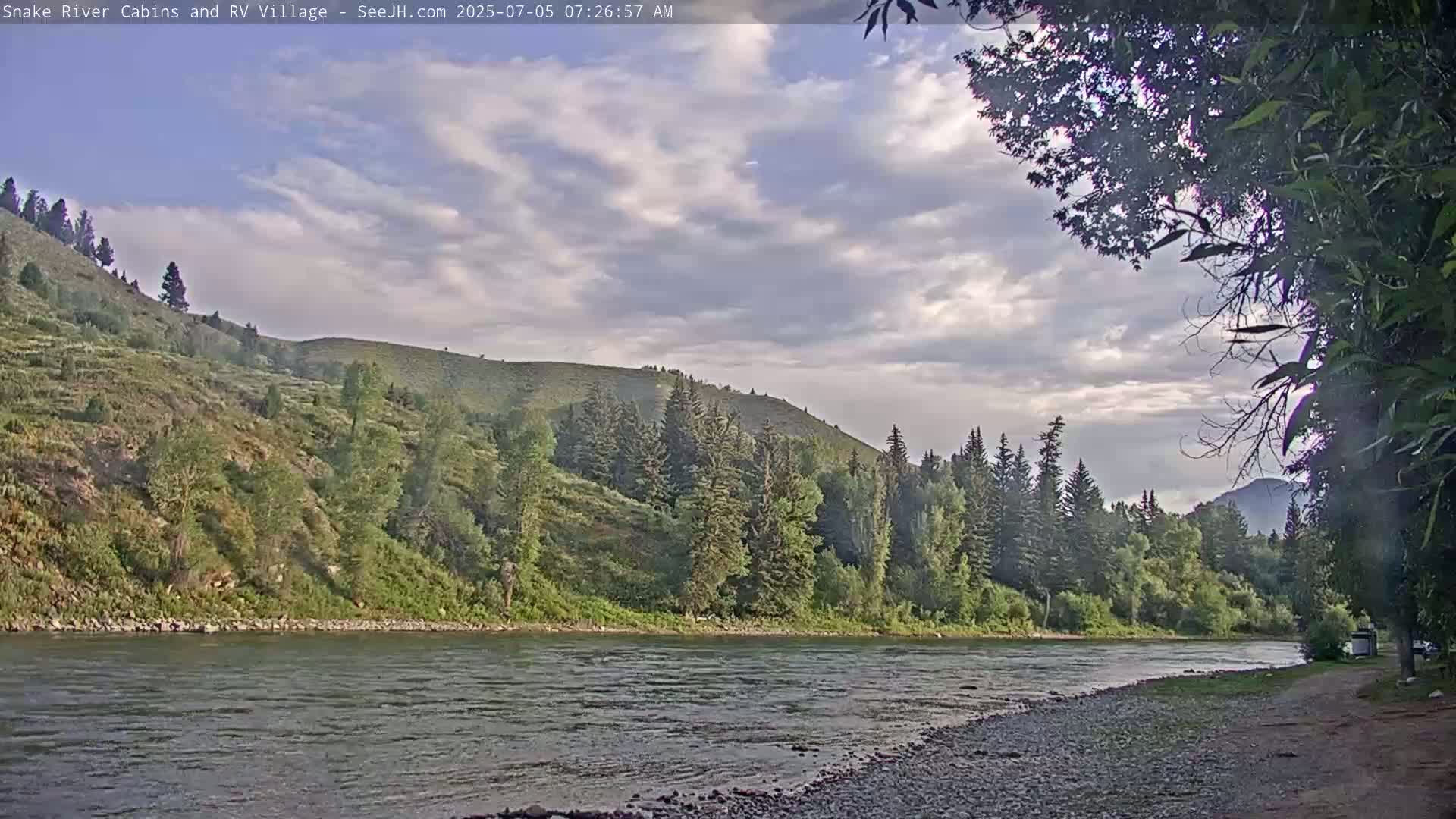 A partly cloudy morning reveals a calm river flowing past a rocky shore and lush green hillside dotted with evergreen trees.