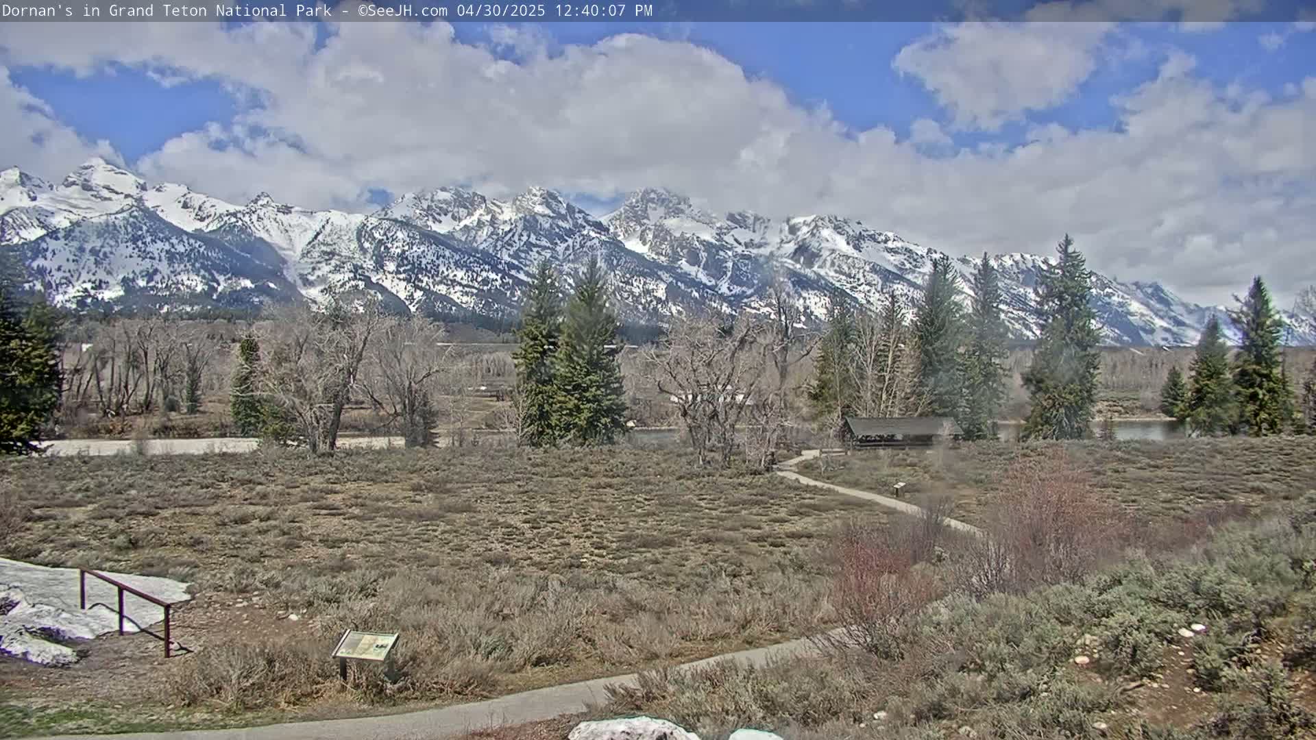 A partly cloudy day reveals snow-capped mountains in the distance, overlooking a flat, scrubby landscape with a few trees, a small structure, and a paved path.