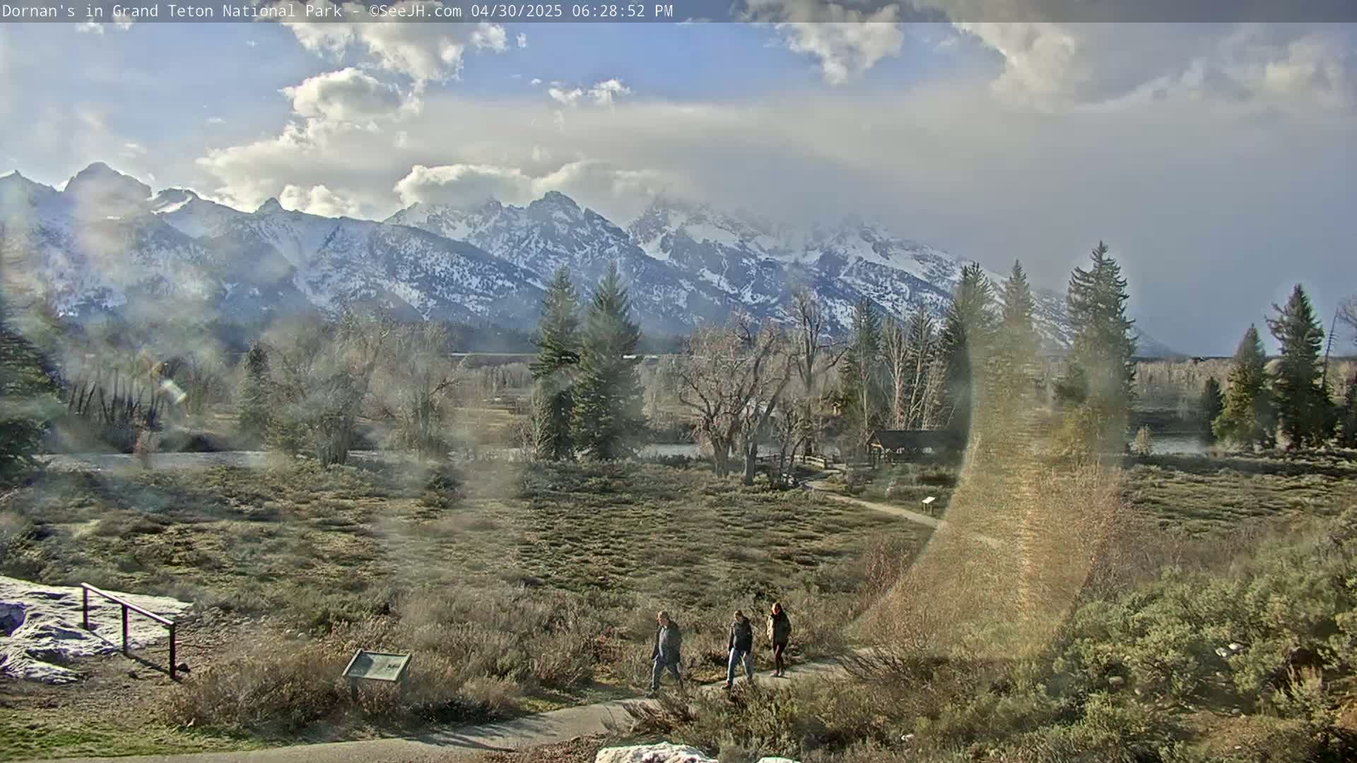 Three people walk on a path through a flat, scrubby area toward snow-capped mountains under a partly cloudy sky.