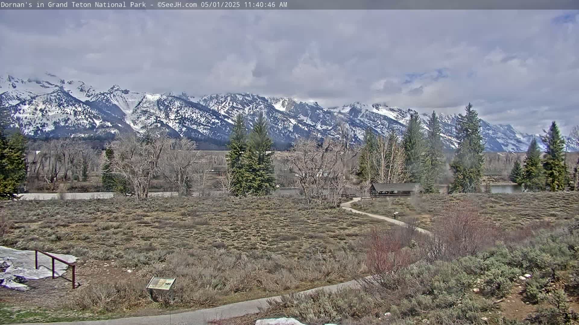 A snow-capped mountain range is visible in the background under a partly cloudy sky, with a flat, sparsely vegetated foreground containing a paved path and a small structure.