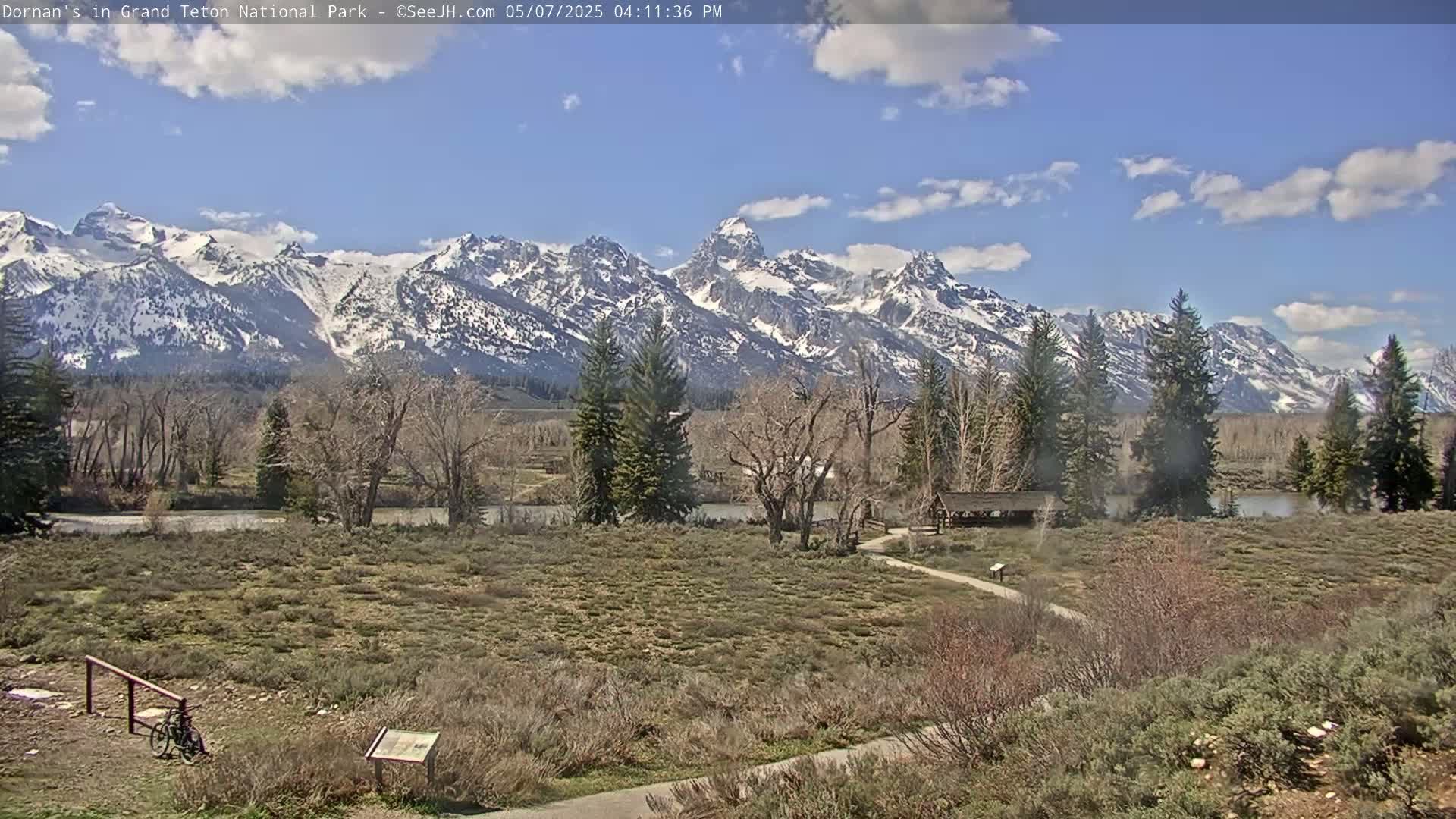 A partly cloudy day shows a snow-capped mountain range in the distance behind a flat expanse of scrubby vegetation, a calm river, and a few leafless trees.