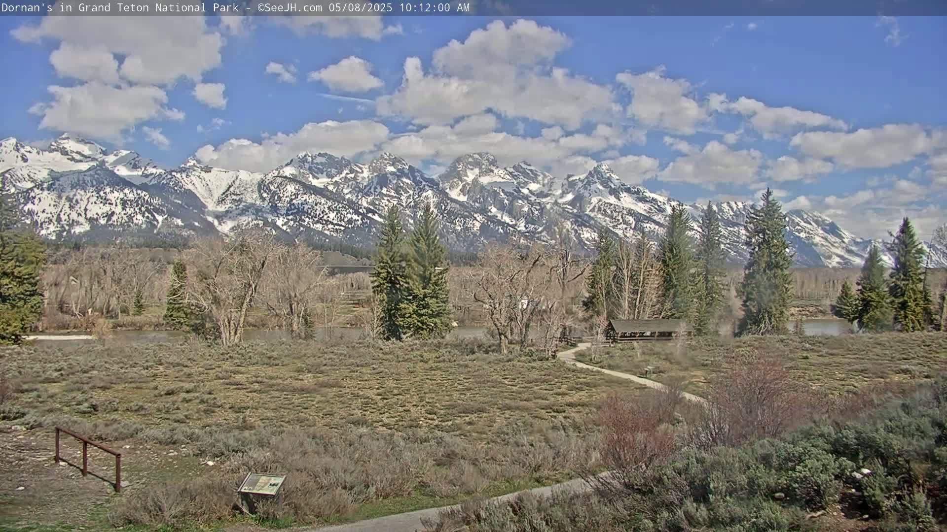 A partly cloudy day reveals a snow-capped mountain range in the background, with a flat, shrubby foreground containing a small structure and pathway near a river.