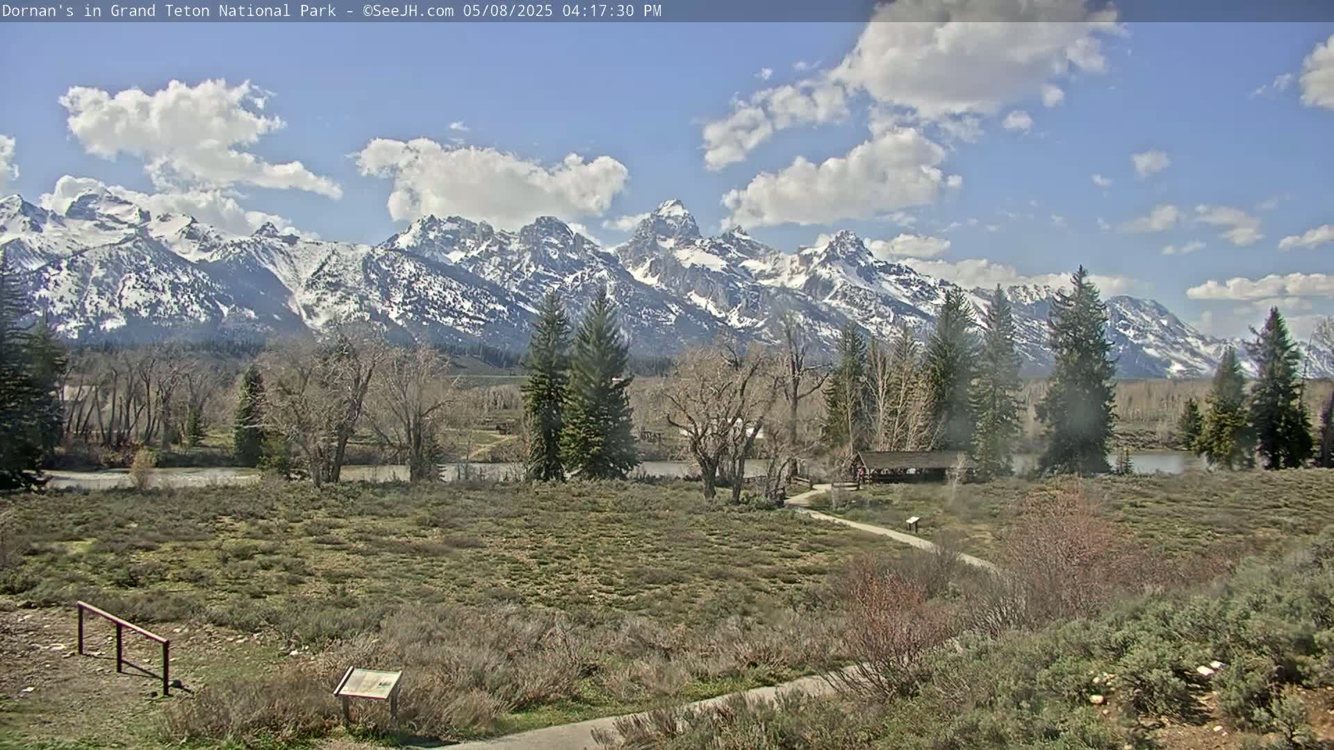 A partly cloudy day reveals snow-capped mountains in the background, with a flat, grassy foreground featuring trees, shrubs, and a paved path leading to a covered structure near a body of water.