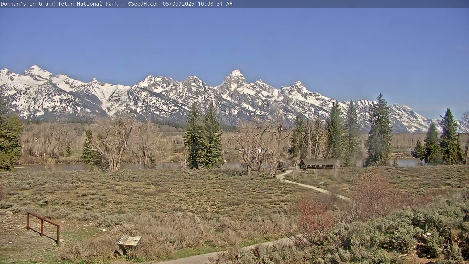 A snow-capped mountain range rises in the background under a clear blue sky over a flat, dry landscape with a river, sparse vegetation, and a small building.