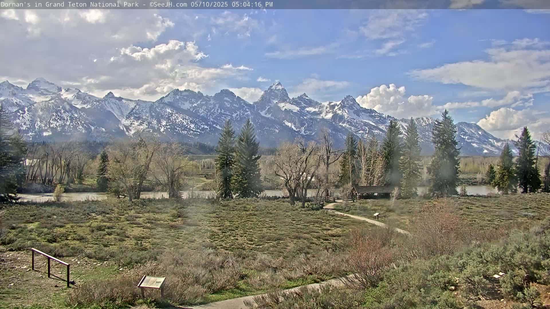 A partly cloudy day reveals a snow-capped mountain range in the distance, overlooking a flat, grassy area with trees and a small structure near a river.