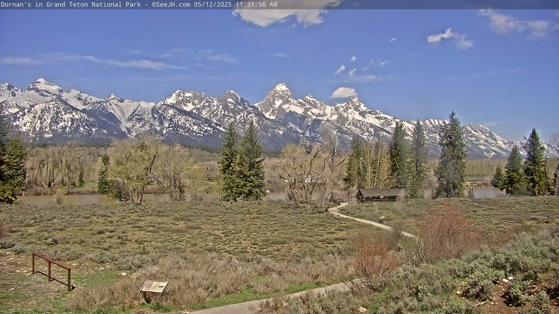 A mostly sunny day reveals snow-capped mountains in the distance, overlooking a flat, scrubby landscape with a river and a small wooden structure near a walking path.