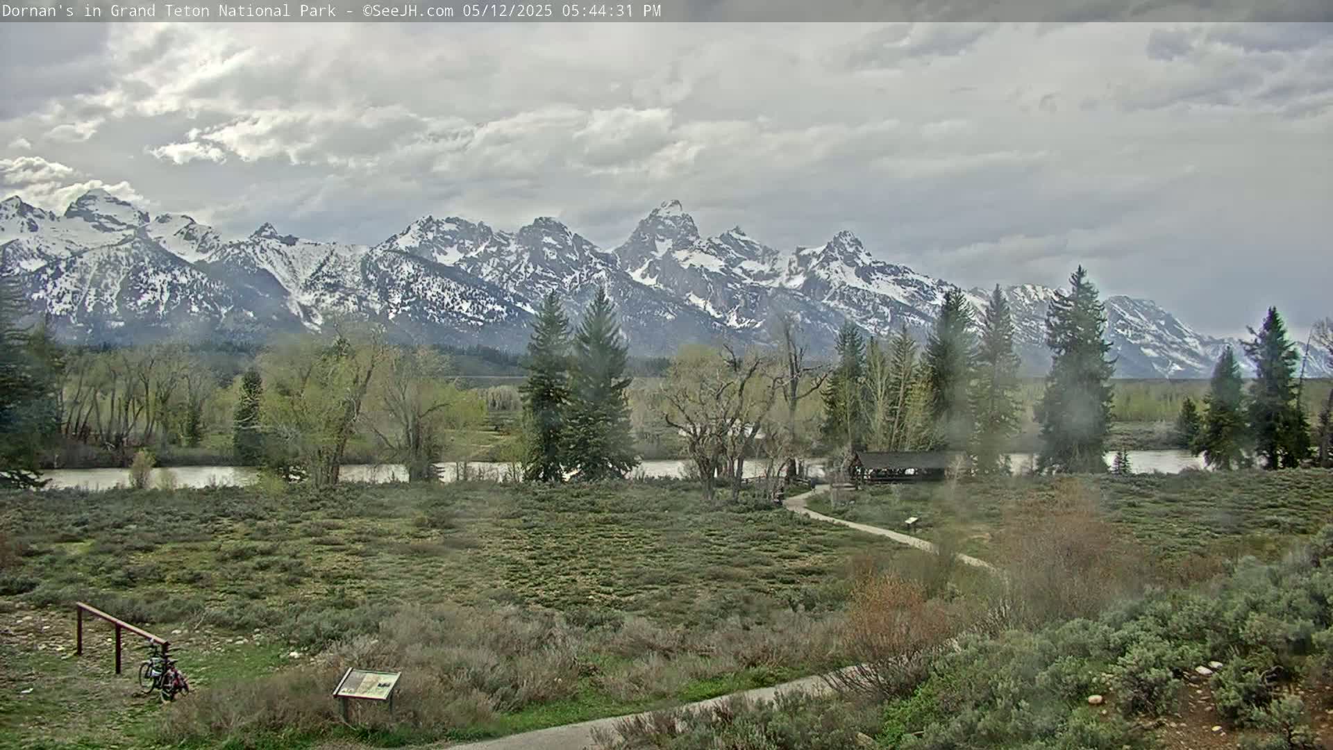 A partially cloudy day reveals a snow-capped mountain range across a flat, green valley with a river and sparse trees, a path, and a bicycle.