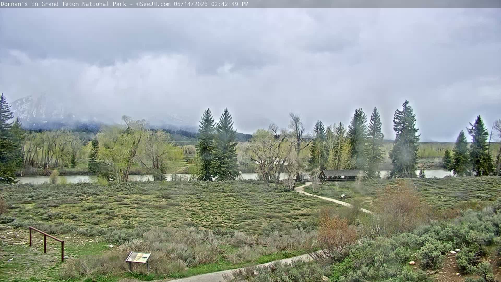 A mostly cloudy day reveals a river flowing through a grassy field with trees and mountains in the background.
