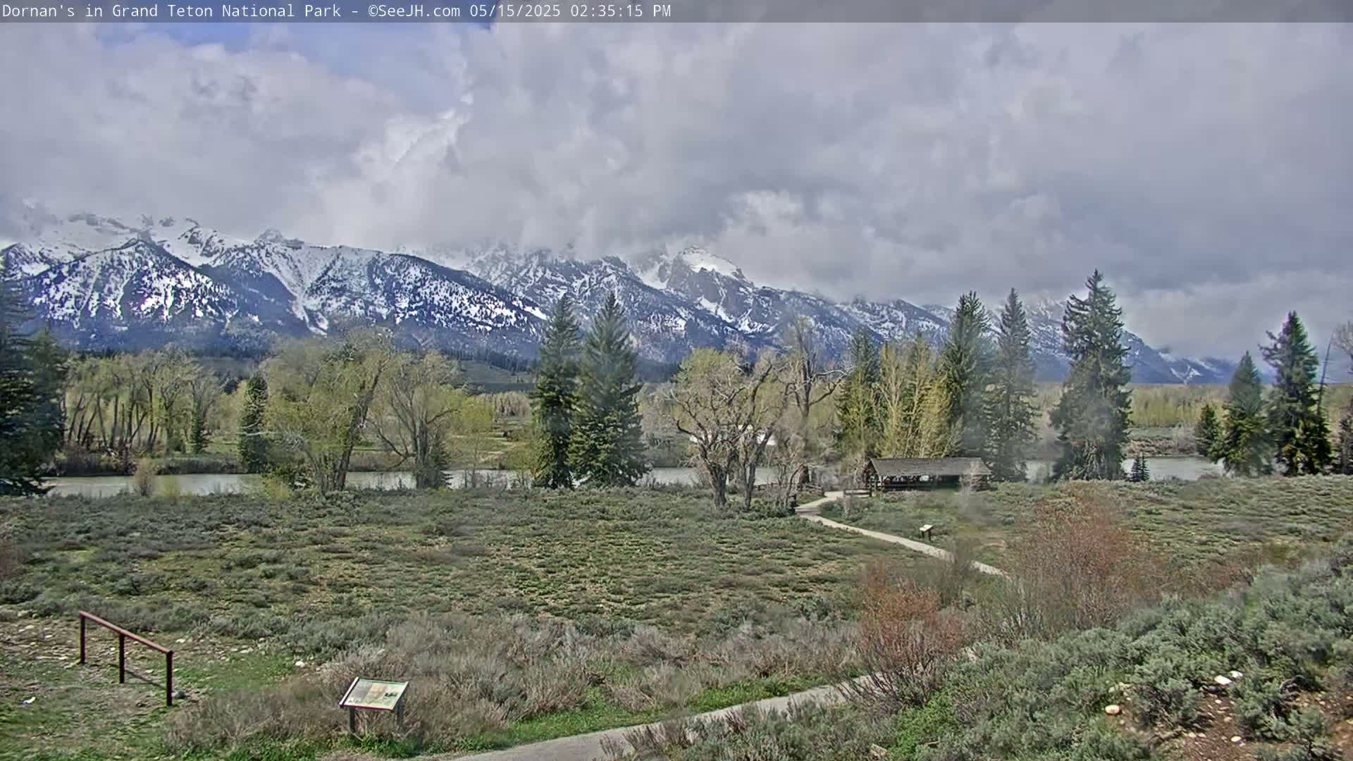 A partly cloudy day shows a river winding through a valley, with snow-capped mountains in the background and budding trees and shrubs throughout the foreground.
