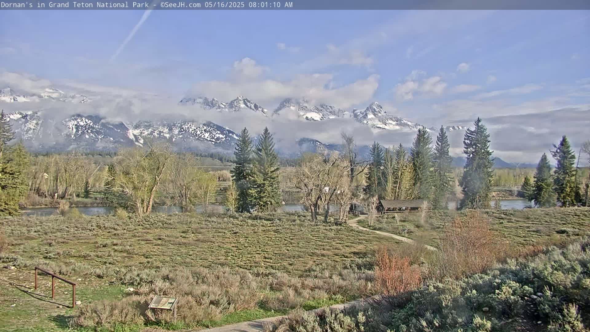 A partly cloudy day reveals snow-capped mountains in the distance, overlooking a flat, brushy landscape with a river, trees, and a small building.