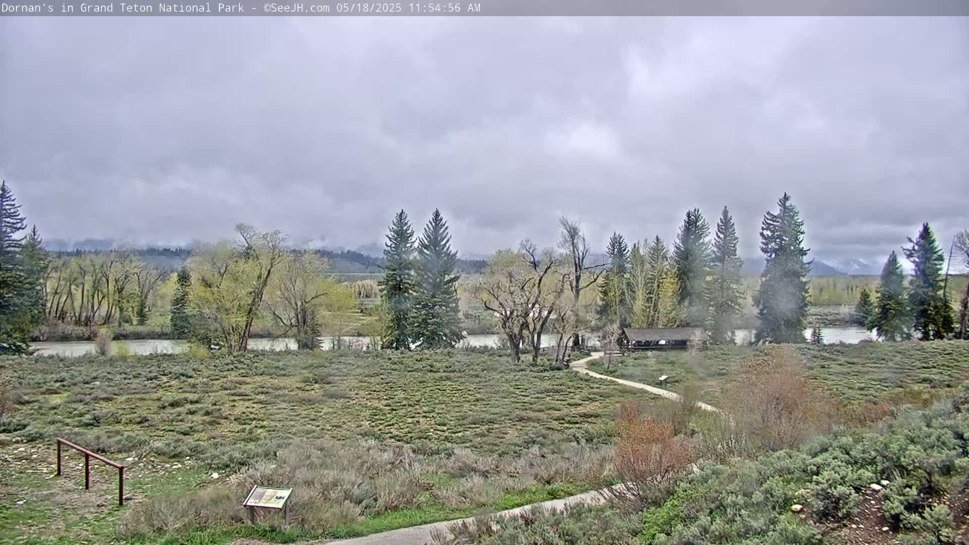 A mostly cloudy day reveals a flat, grassy area with a river, trees, and a small building, all set against a backdrop of distant mountains.