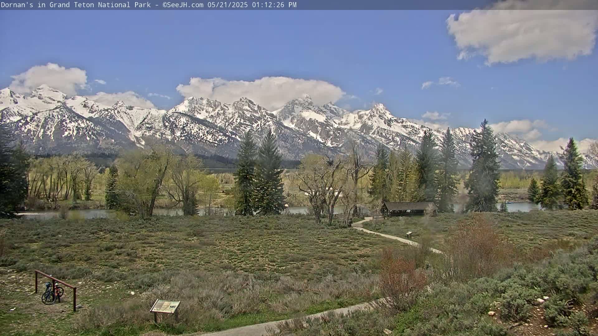 A partly cloudy day reveals snow-capped mountains in the background, with a flat foreground of shrubs and trees bordering a calm river.